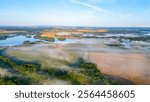 The hazy morning light casts a serene atmosphere over Rozmberk Pond, near Trebon, Czechia. Mist hovers above the water, creating a peaceful landscape with lush greenery and distant fields.