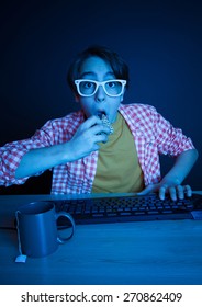 He eat cake while play on computer in the night. In blue light of display emotional kid play computer games online.