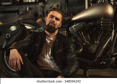 He has character. Portrait of a handsome sexy bearded guy looking to the camera confidently while relaxing near his motorcycle at the custom workshop lifestyle garage relax hobby concept