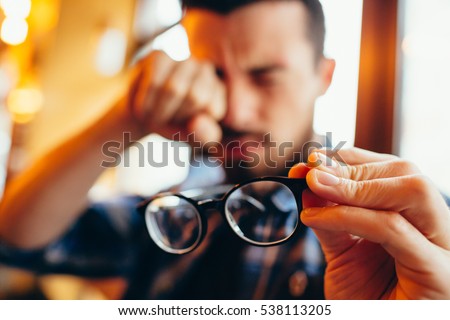 He has eyesight problems. guy with beard is holding his eyeglasses right in front of camera with one hand and rubbing his eyes with another hand. Focus is on eyeglasses