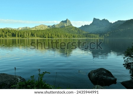 he rocky shore of a large lake in the mountains with a reflection of a dense forest and high rocks in the unsteady surface of the water on a sunny summer evening. Lake Svetloe, Ergaki Nature Park, 