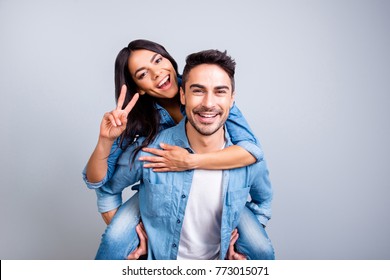 he and she travel together, love story of attractive, happy, smiling, sweet couple in casual outfit, man carrying his lover on back, woman showing peace symbol over grey background - Shutterstock ID 773015071