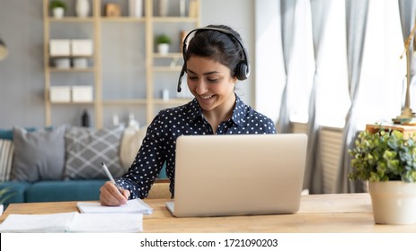 Head Ahot Happy Indian Girl Student Wearing Headphones, Watching Educational Lecture Workshop Seminar, Writing Notes. Smiling Hindu Woman Studying Online, Sitting At Desk With Computer At Home.