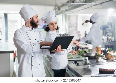 Head chef with modern laptop showing to sous chef recipe for gourmet dish required for gastronomic event held at restaurant. Food industry workers standing in professional kitchen while using computer - Powered by Shutterstock