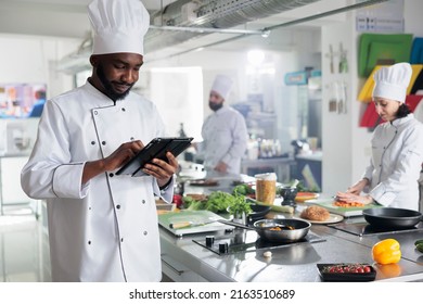 Head chef with modern touchscreen tablet device searching for gourmet cuisine dish recipe while colleagues chopping fresh vegetables. Gastronomy expert with handheld device standing in kitchen. - Powered by Shutterstock