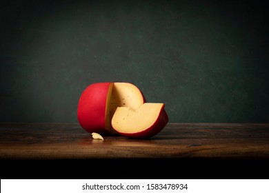 Head Of Edam Cheese With A Red Crust Lying On A Brouwn Wooden Table With A Green Background. Rembrandt Lightning Style