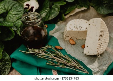 A Head Of Goat's Milk Cheese With Spices With A Jar Of Honey, Almond Nuts And A Sprig Of Rosemary On A Green Background.