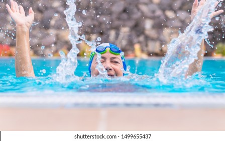 Head Of An Old Lady Peeks Out Of The Pool Water. Healthy Pool Activity. Partially Hidden By The Water Spray. With Cap And Googles. People Smiling And Playing Sports