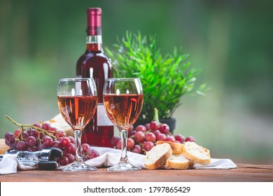 Head On Shot Of Two Glasses Of Rose Wine Outside On A Rustic Wooden Picnic Table Surrounded By Fresh Red Grapes, French Bread, And A Wine Bottle.