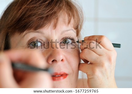 Head portrait in the mirror of a brunette middle-aged woman pulling an eyeliner with a pencil in front of the mirror.