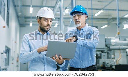 Head of the Project Holds Laptop and  Discusses Product Details with Chief Engineer while They Walk Through Modern Factory.