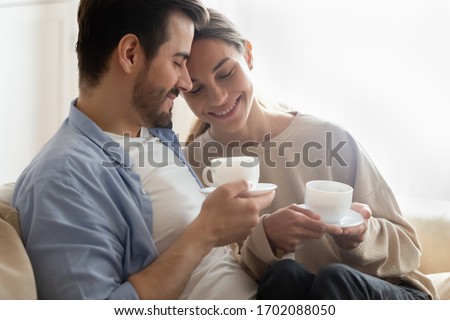 Head shot affectionate loving married couple bonding, drinking hot coffee, enjoying spending morning time together at home. Emotional spouses relaxing on comfortable couch, sweet tender moment.