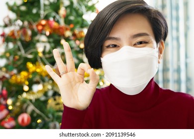 Head Shot Of A Beautiful Asian Woman In Red With Medical Face Mask Happy Smiling And Do Okay Hand Sign To Her Friends And Family Through Web Cam Video Call In Christmas Eve While She's Quarantine.