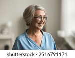 Head shot cheerful pretty elderly woman with short, grey hair standing indoor looking away, wearing eyeglasses and blue shirt posing alone in living room, smile, reflecting, deep in pleasant memories