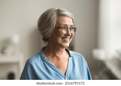 Head shot cheerful pretty elderly woman with short, grey hair standing indoor looking away, wearing eyeglasses and blue shirt posing alone in living room, smile, reflecting, deep in pleasant memories - Powered by Shutterstock