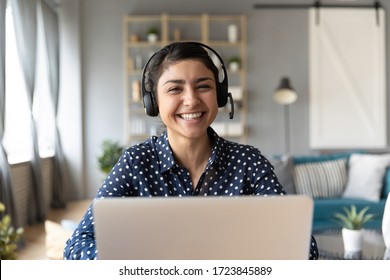 Head Shot Cheerful Smiling Pretty Hindu Ethnic Girl Sitting At Table With Computer, Wearing Headphones With Mic, Looking At Camera. Happy Indian Woman Professional Tutor Educating Client Online.