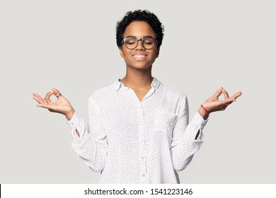 Head shot close up calm African American young woman in glasses meditating, beautiful girl with closed eyes and toothy smile practicing yoga, stress relief concept, isolated on grey background