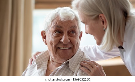 Head shot close up cheerful elderly man looking at pleasant middle aged nurse. Mature female doctor embracing shoulders, communicating with smiling 80s patient, giving support and psychological help.