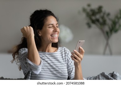 Head shot close up of excited woman holding smartphone, celebrating success, online lottery win, looking at phone screen, overjoyed young female rejoicing, showing yes gesture, received good news