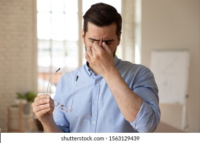 Head shot close up exhausted young businessman taking off eyeglasses, massaging nose bridge. Tires stressed overworked male manager worker employee suffering from dry eyes syndrome or eyestrain.