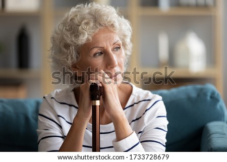 Head shot close up front view pondering stressed woman sitting on couch with wooden walking stick, thinking of health problem alone at home. Unhappy depressed grandmother suffering from disability.