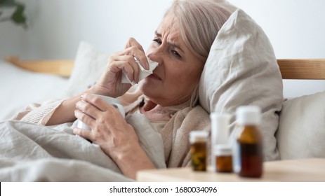 Head Shot Close Up Ill Mature Woman Lying In Bed, Using Paper Tissue, Wiping Runny Nose. Unhealthy Middle Aged Retired Lady Caught Cold, Drinking Hot Tea, Taking Antibiotic Pills From Bedside Table.