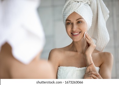 Head Shot Close Up Mirror Reflection Smiling Beautiful Woman Wrapped In Towel Applying Toner With Cotton Pad On Skin Face After Bath. Happy Young Lady Doing Morning Skincare Routine After Shower.