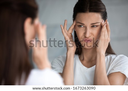 Head shot close up mirror reflection unhappy young brunette woman touching eyes area, feeling dissatisfied with first wrinkles, ineffective skincare products, confused with bad skin condition.