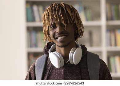 Head shot close up portrait African guy with dreadlock hairstyle, headphones on neck and backpack smile look at camera pose in library on bookshelves background. Education, studentship, new knowledge - Powered by Shutterstock