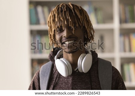 Head shot close up portrait African guy with dreadlock hairstyle, headphones on neck and backpack smile look at camera pose in library on bookshelves background. Education, studentship, new knowledge