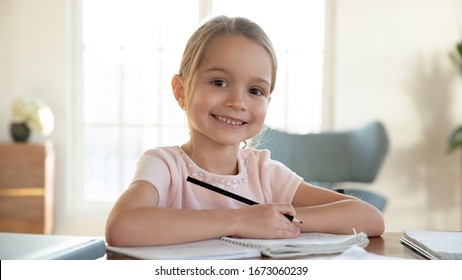 Head shot close up portrait of happy small pupil learning at home. Smiling little child girl enjoying doing lessons in living room. Smart kid schoolgirl looking at camera, studying remotely online.