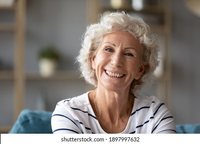 Head shot close up portrait of happy grey-haired old retired woman sitting on sofa. Sincere smiling middle aged grandmother enjoying video call conversation with friends or grown children, screen view
