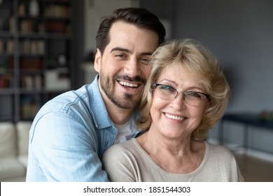 Head shot close up portrait of loving young bearded man cuddling smiling beautiful middle aged mature mother on eyeglasses. Loving different generations family looking at camera, enjoying weekend.