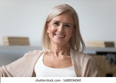 Head shot close up portrait of pleasant smiling mature woman. Happy healthy middle aged lady relaxing alone at home, looking at camera, posing for photo. Positive satisfied retirement concept.