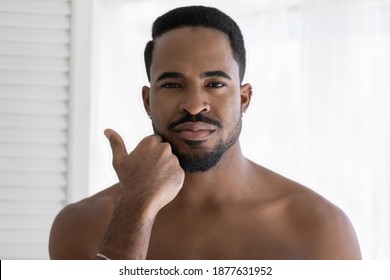 Head shot close up portrait of young african ethnicity man touching beard, satisfied with daily shaving routine. Handsome millennial shirtless mixed race guy feeling confident, looking at camera.