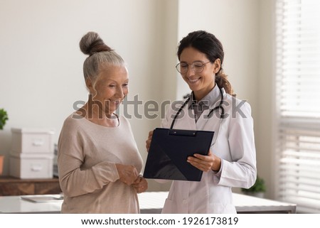 Head shot close up smiling female doctor wearing glasses and white uniform consulting mature patient about treatment, holding clipboard, happy senior woman and physician standing in hospital