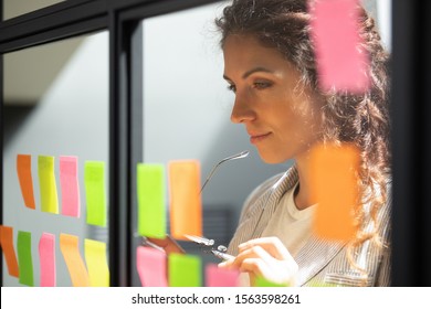 Head shot close up thoughtful young female boss team leader looking at kanban scrum glass window board, developing start up project marketing strategy, managing workflow, checking tasks at office.