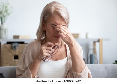 Head shot close up tired older lady taking off eyeglasses, suffering from dry eyes syndrome. Unhealthy middle aged granny massaging nose bridge, reducing eyes tension or strain, sitting on sofa. - Powered by Shutterstock