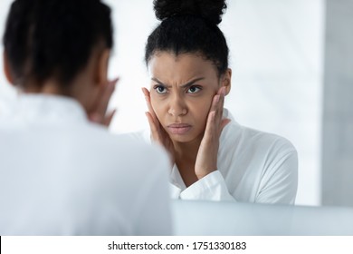 Head Shot Close Up Unhappy African American Young Woman Checking Skin, Touching Cheeks, Angry Girl Wearing White Bathrobe Worried About Mimic Wrinkle Or Acne, Looking In Mirror, Standing In Bathroom