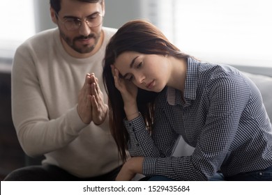 Head shot close up upset abused offended woman sitting on sofa, ignoring her apologizing husband. Young man feeling sorry, asking forgiveness to depressed wife. Family relationship problems concept.
