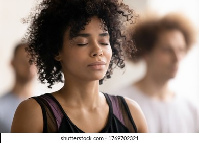 Head shot close up young peaceful attractive curly hair african american woman breathing fresh air, enjoying deep meditation with closed eyes, relaxing after yoga class workout in sport club.