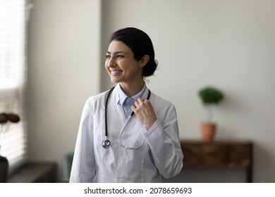 Head Shot Dreamy Smiling Indian Female Doctor Therapist Nurse In White Uniform With Stethoscope Looking In Distance, Thinking About Success, New Job Opportunities, Standing In Medical Office