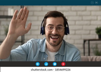 Head shot emotional happy 30s man in eyeglasses and wireless headphones, looking at camera, making hello gesture, greeting friends starting online video call, computer application monitor view.