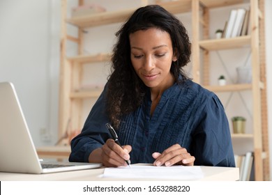 Head Shot Focused Young Mixed Race Female Manager Company Worker Sitting At Desk Alone In Office, Working With Financial Documents. Concentrated Professional Writing Notes While Watching Webinar.