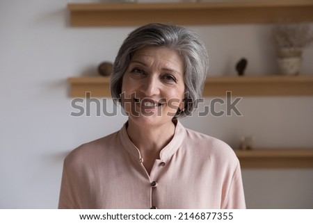Head shot of good-looking elegant middle-aged woman having charming smile posing standing alone indoor staring at camera. Professional tutor portrait, medical insurance cover for older citizen concept