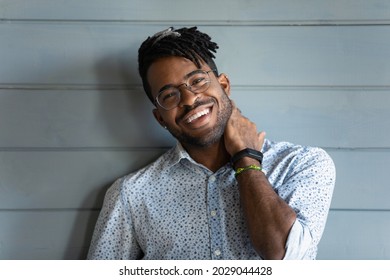 Head Shot Of Happy Black Mixed Race Guy With Trendy Haircut, Glasses, Toothy Smile, Wearing Stylish Casual Clothes, Touching Stubble, Neck Skin, Laughing At Camera. Handsome Millennial Man Portrait