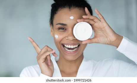 Head shot of happy excited mixed race woman in bathrobe with moisturizing cream spots on face covering eye with cosmetic jar circle, pointing finger away, showing copy space. Close up of face - Powered by Shutterstock