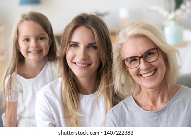 Head shot of happy feminine three generations indoors. Portrait of beautiful little granddaughter daughter and grandmother smiling looking at camera sitting together on couch in living room at home
