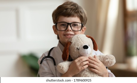 Head shot happy little cute kid boy in glasses and medical coat cuddling teddy bear toy, pretending to be veterinary doctor. Smiling small smart child looking at camera, playing with fluffy toy.