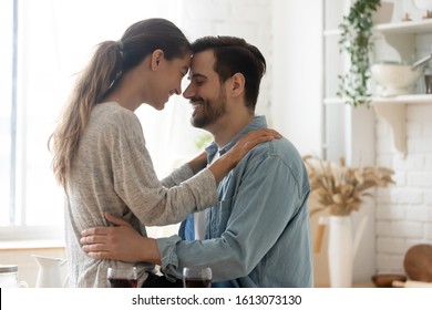 Head shot happy loving mixed race couple touching foreheads, enjoying sweet tender moment in kitchen. Smiling young man cuddling sitting on countertop wife, spending free time together at home.